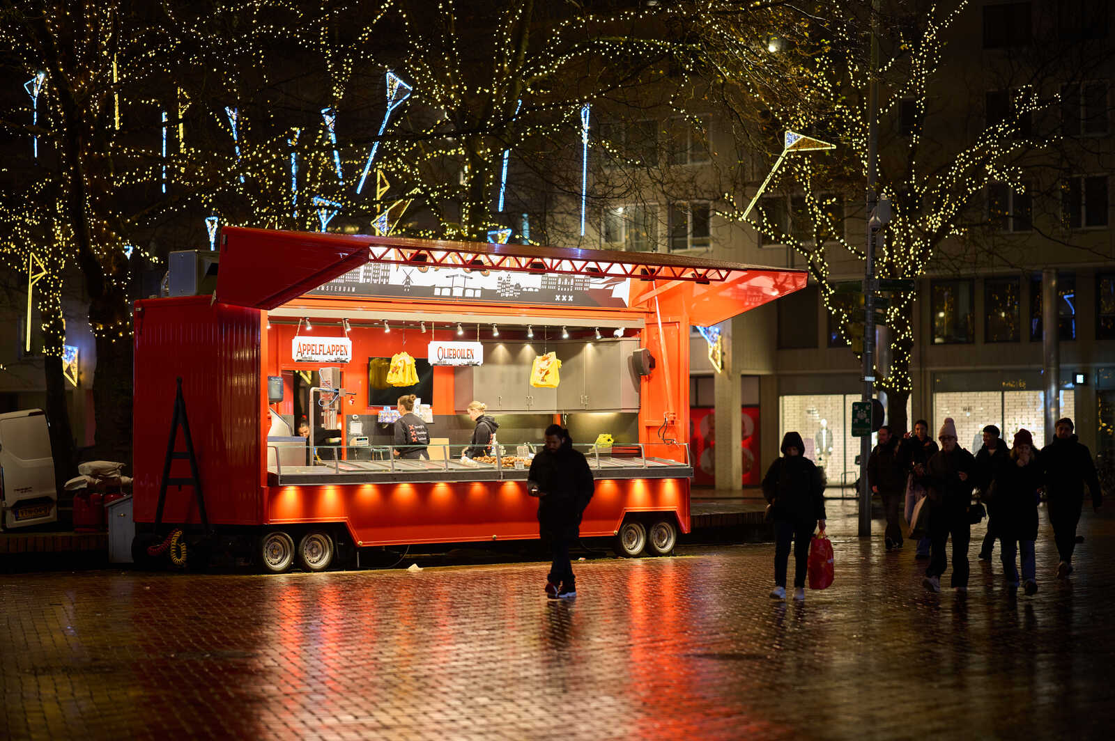 A red food cart displaying pastries under warm lights. People walk by on a wet pavement at night, with string lights overhead. The cart has Appelflappen and Oliebollen signs.