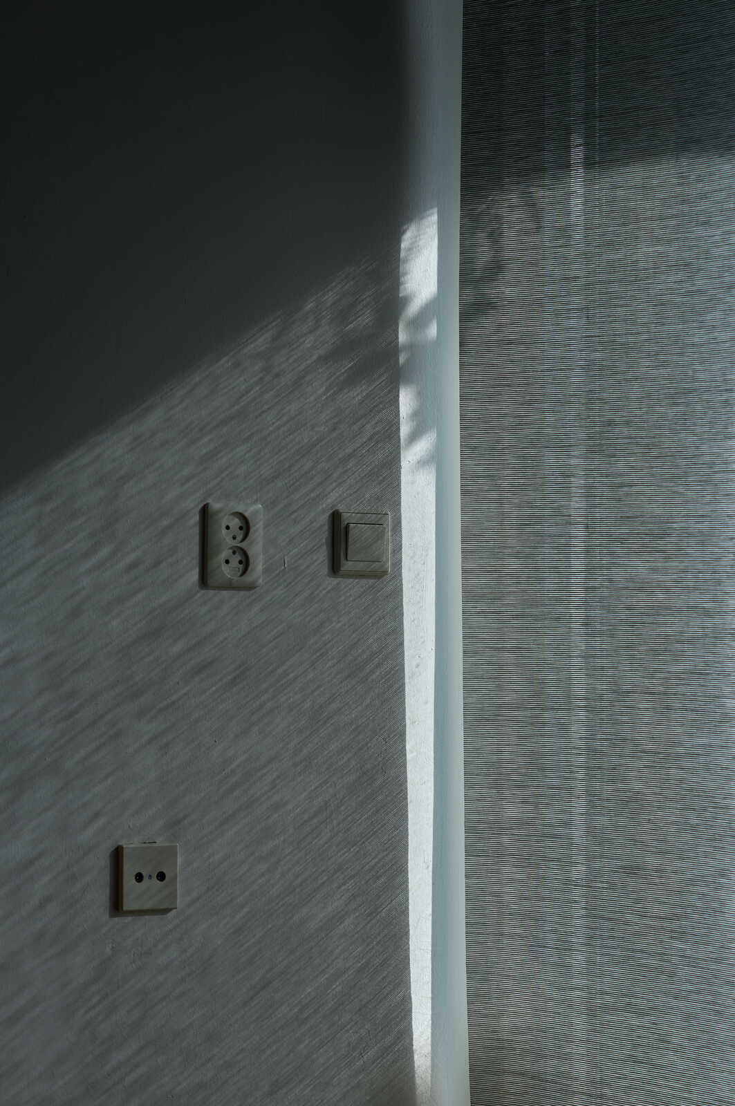 Three gray electrical outlets are affixed to a textured, pale gray wall. A corner of the wall is visible on the left. The wall's texture runs vertically.