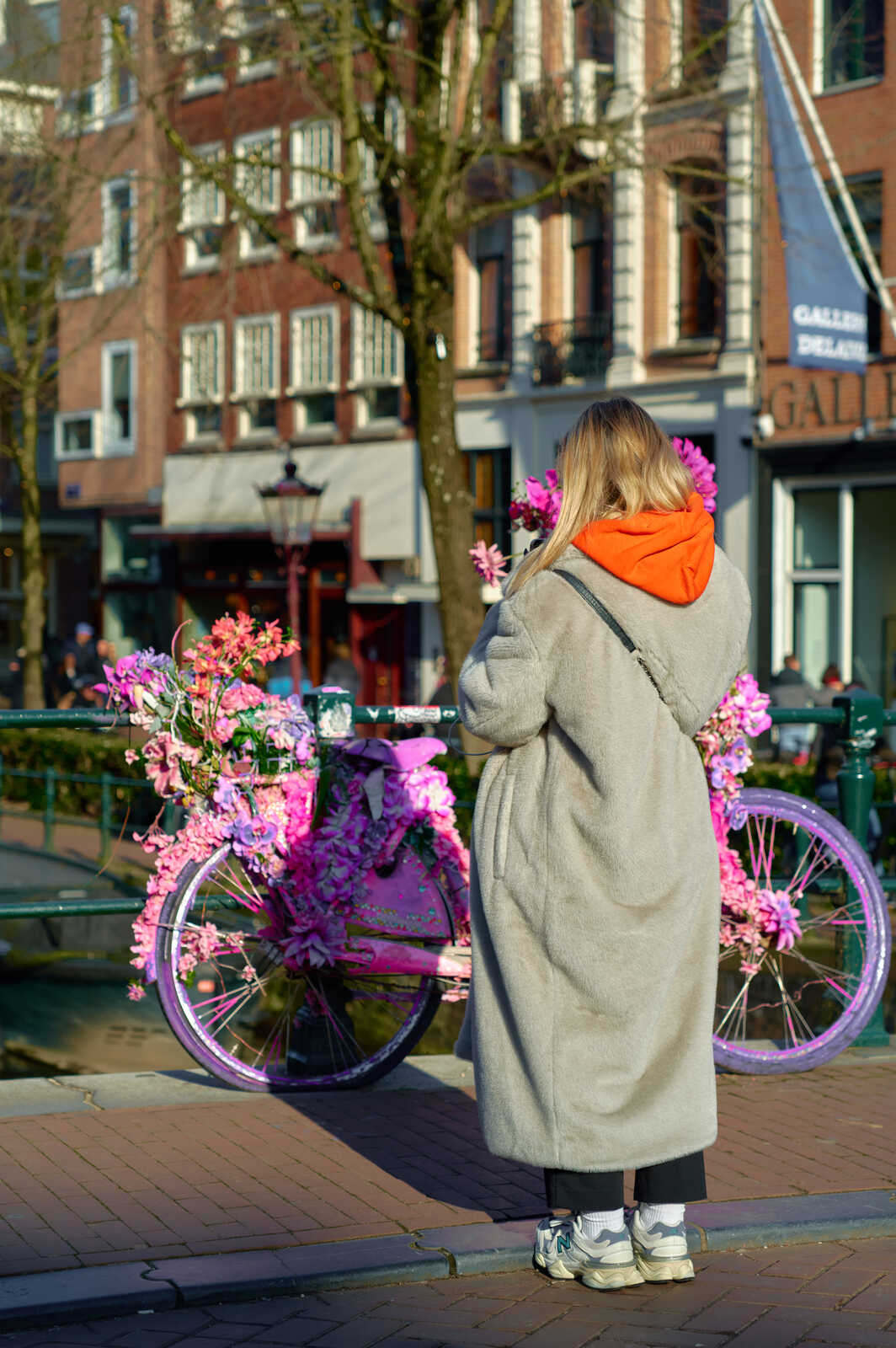 A person in a gray coat stands next to a bicycle adorned with pink flowers. A canal and building are in the background. A sign reads Galerie.
