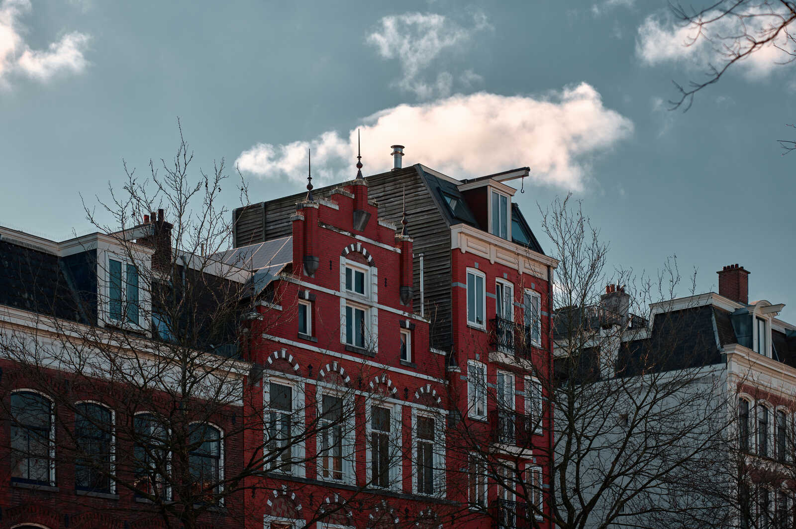 Red brick building with white decorative trim, a steeply pitched roof, dormer windows, and a few chimneys. Bare tree branches in the foreground. Cloudy sky above.