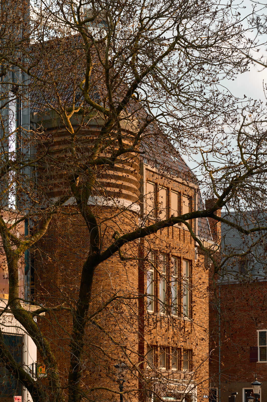 A brick building with a curved facade and arched windows is visible, partially obscured by bare tree branches against a cloudy sky. A modern glass building is seen to the left.