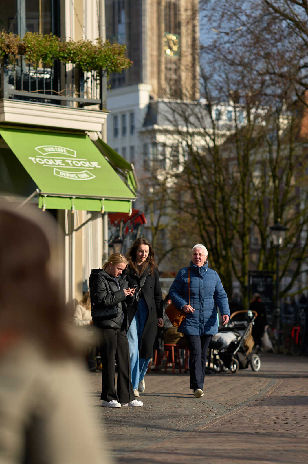 Two people walk on a city street. One has a dark jacket and looks at their phone. The other has a blue jacket and carries a blue bag. A cafe is visible on the left with a green awning.