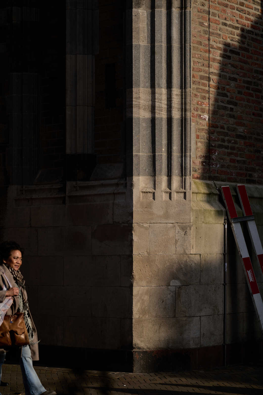 A person in jeans and a scarf walks past a brick building with a metal ladder leaning against it. The building has a stone facade with a window. The sidewalk is visible.