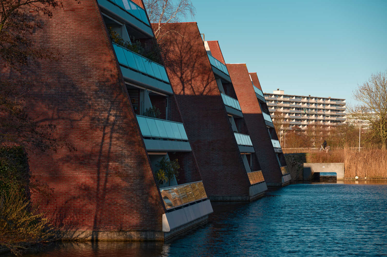 Red brick buildings with angled balconies face a calm waterway. A person walks on a path behind the water, with a larger building in the distance. Green foliage is on the left.