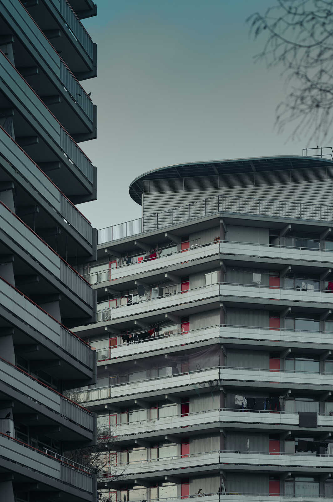 Gray apartment building with red balcony doors and railings, multiple stories high. Several balconies have laundry hanging. A round structure is visible on top. Gray sky in the background.