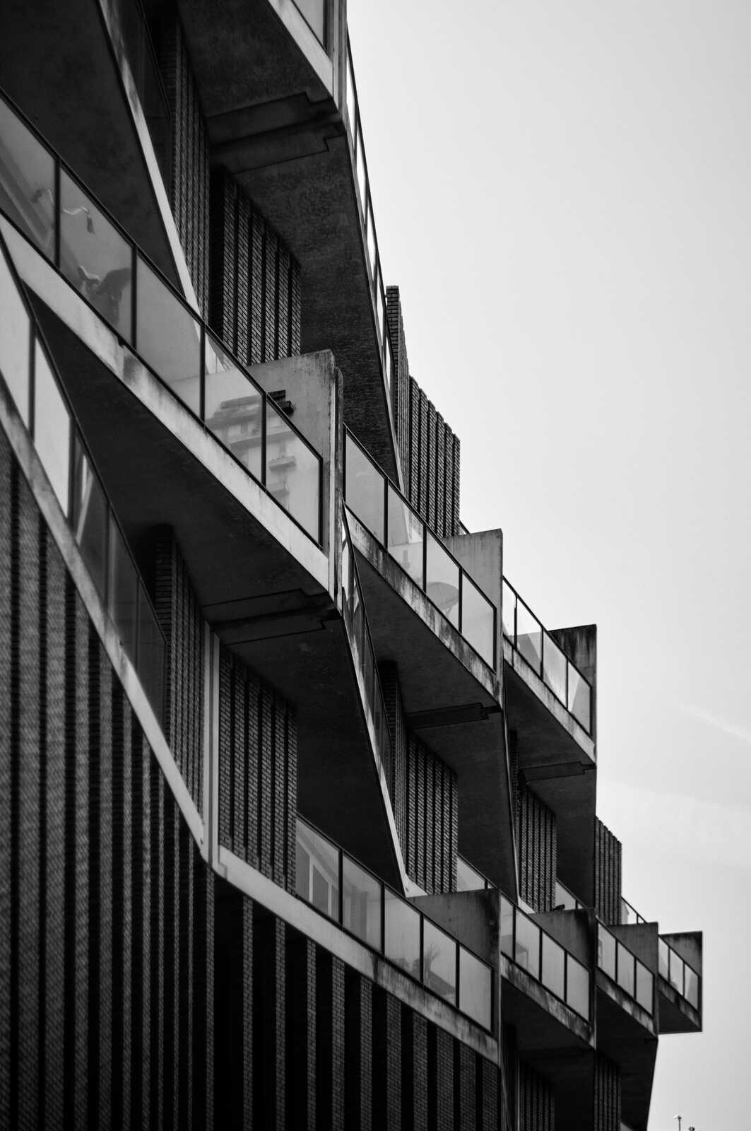 A black and white photo depicts a buildings facade with staggered, cantilevered balconies. Concrete slabs form the structure, with glass panels filling the balcony spaces. Vertical lines accentuate the design.