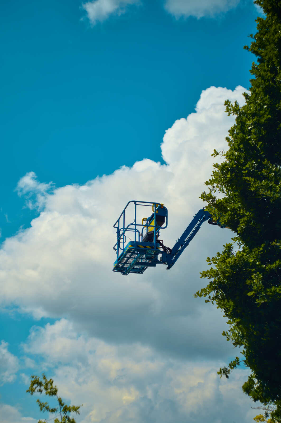 A scissor lift with a person inside is positioned against a blue sky with fluffy white clouds. Green tree foliage is visible on the left side of the frame.