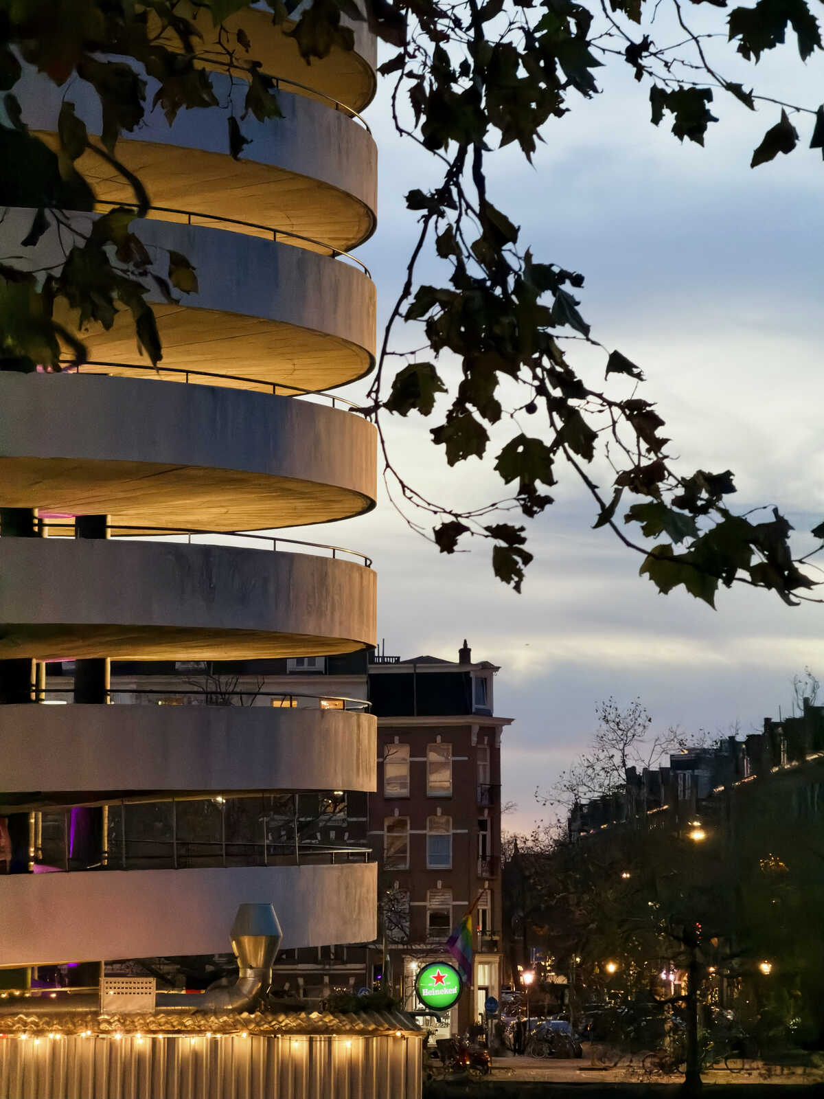 Multi-level parking structure with circular tiers. Warm lights illuminate the structure against a dusky sky. Trees and buildings with lights are visible in the background. A beer sign is present.