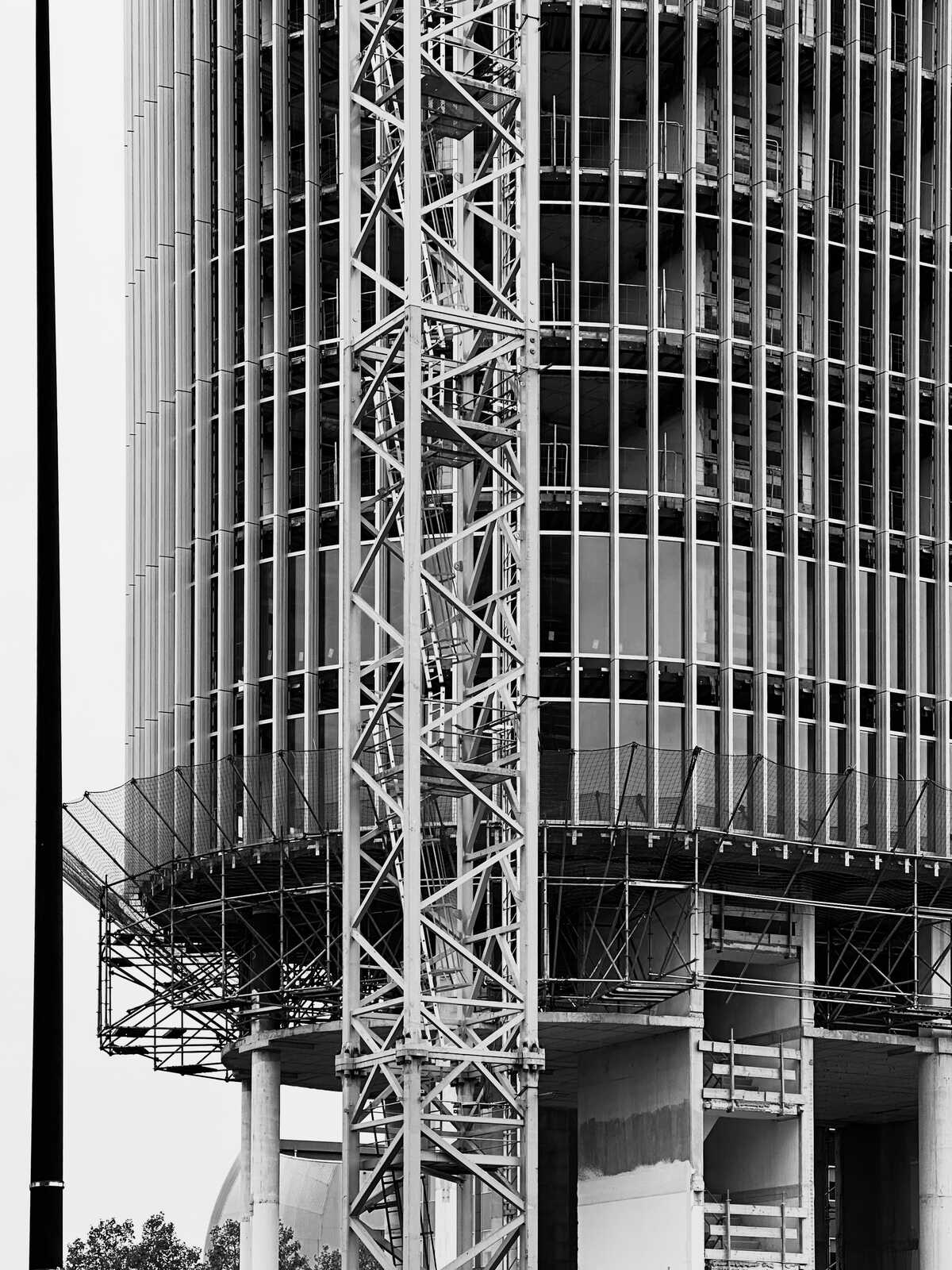 A close-up, grayscale image of a buildings facade. It features vertical metal bars and a grid-like pattern, with a concrete structure visible above. A partial view of a dome is seen in the background.