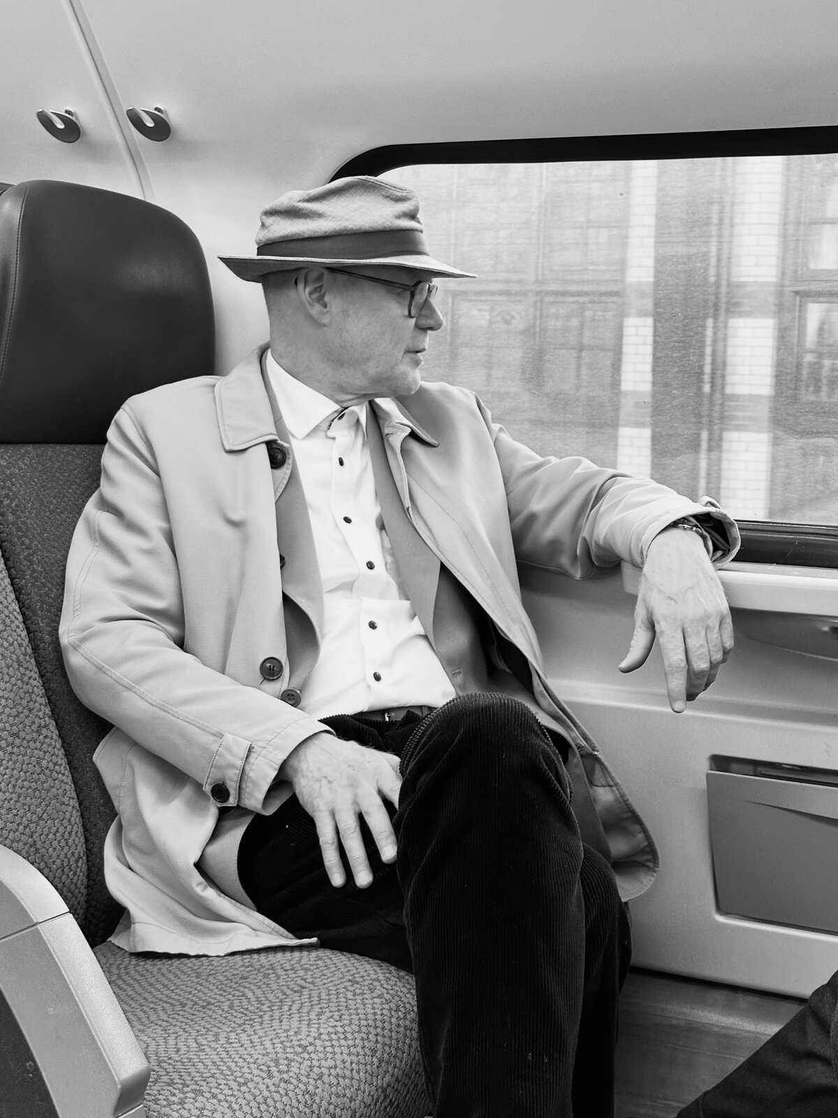 A man in a hat, light-colored coat, and corduroy pants sits in a seat. He has a white shirt and his hand rests on his thigh. A window is behind him.