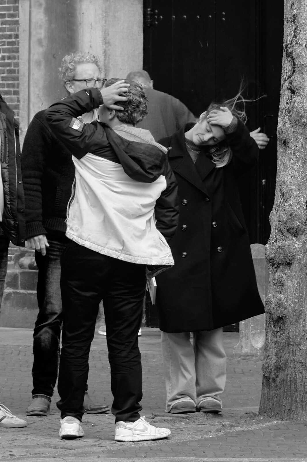 Two people embrace outside a building with a black door. One wears a black coat and the other a white jacket and dark pants. Both have shoes on. A third person is partially visible.