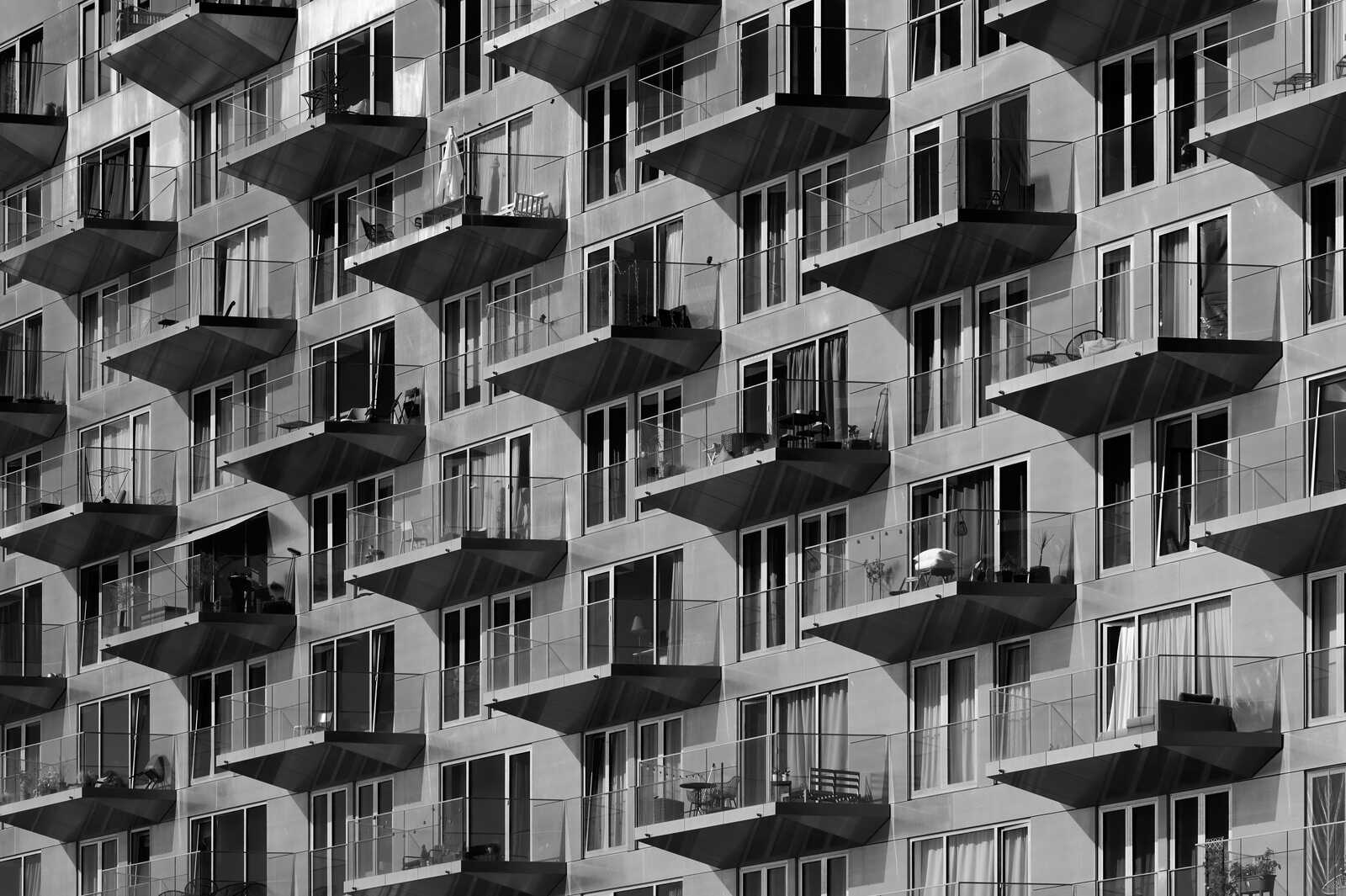 Black and white image of a buildings facade with numerous balconies. Each balcony features glass railings and varying depths. Some have furniture, others are empty. A repeating pattern creates a textured, geometric design.