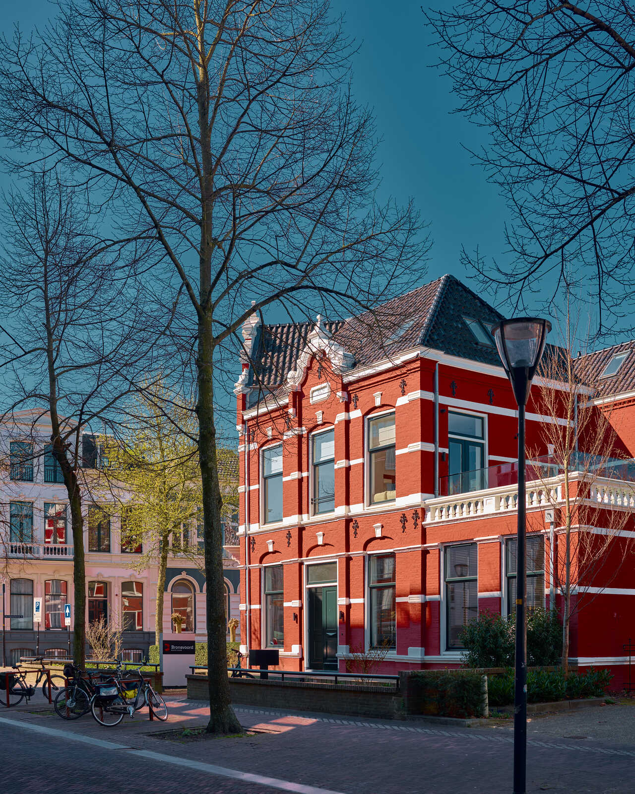 Red brick building with white trim and bay windows. A tree stands prominently in front. Theres a bench and bikes visible on the sidewalk. Blue sky in background.