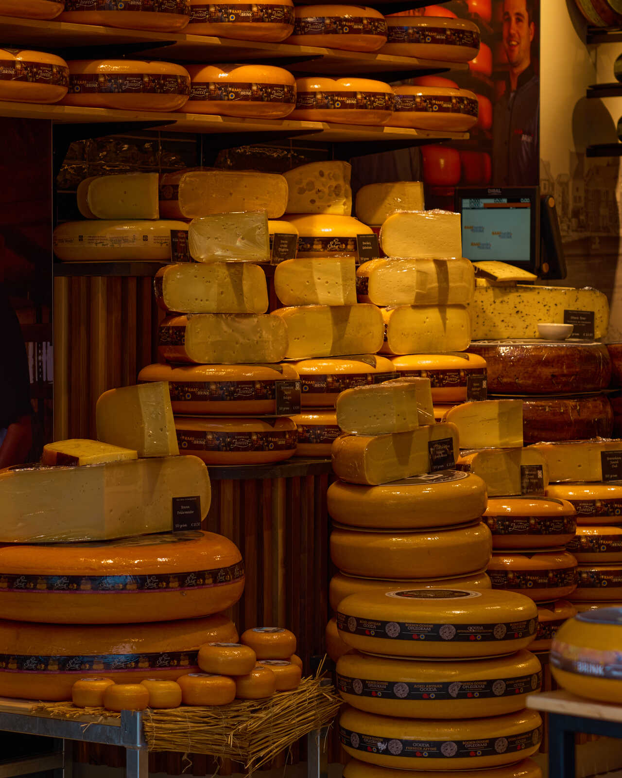 Rows of large, round cheeses with orange wax paper coverings are stacked high on shelves. A man stands behind the display in a dark apron. Signs label the cheeses. A rope lies on a shelf to the right.