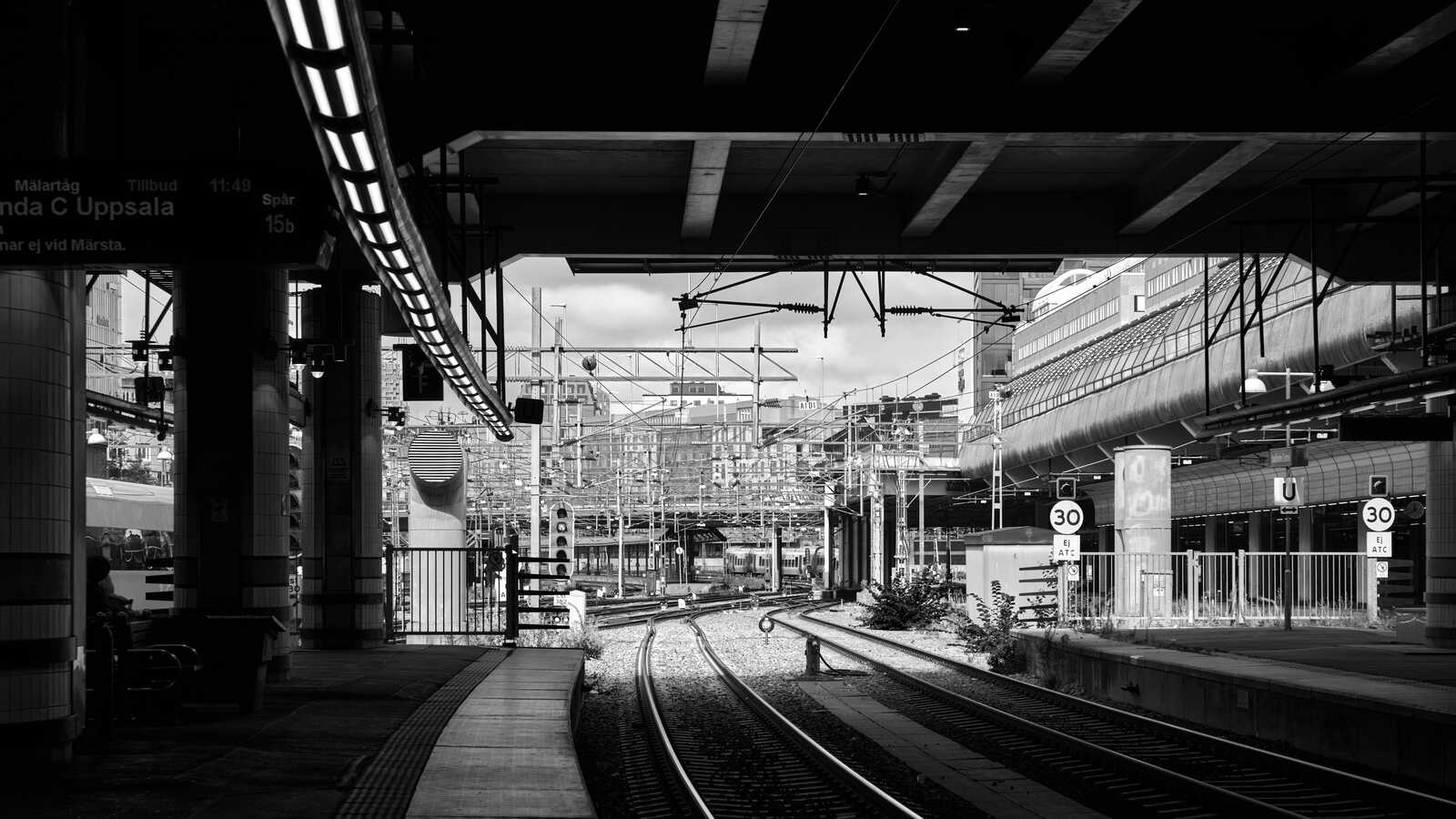 Black and white photo of train tracks and overhead power lines. A platform with a sign is visible on the left. A sign reads 30.