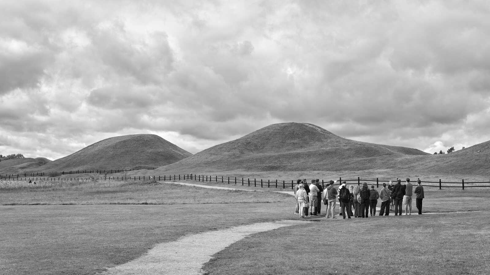 Two grassy hills rise on a field. A path leads to a black fence and a group of people standing nearby. The sky is overcast.
