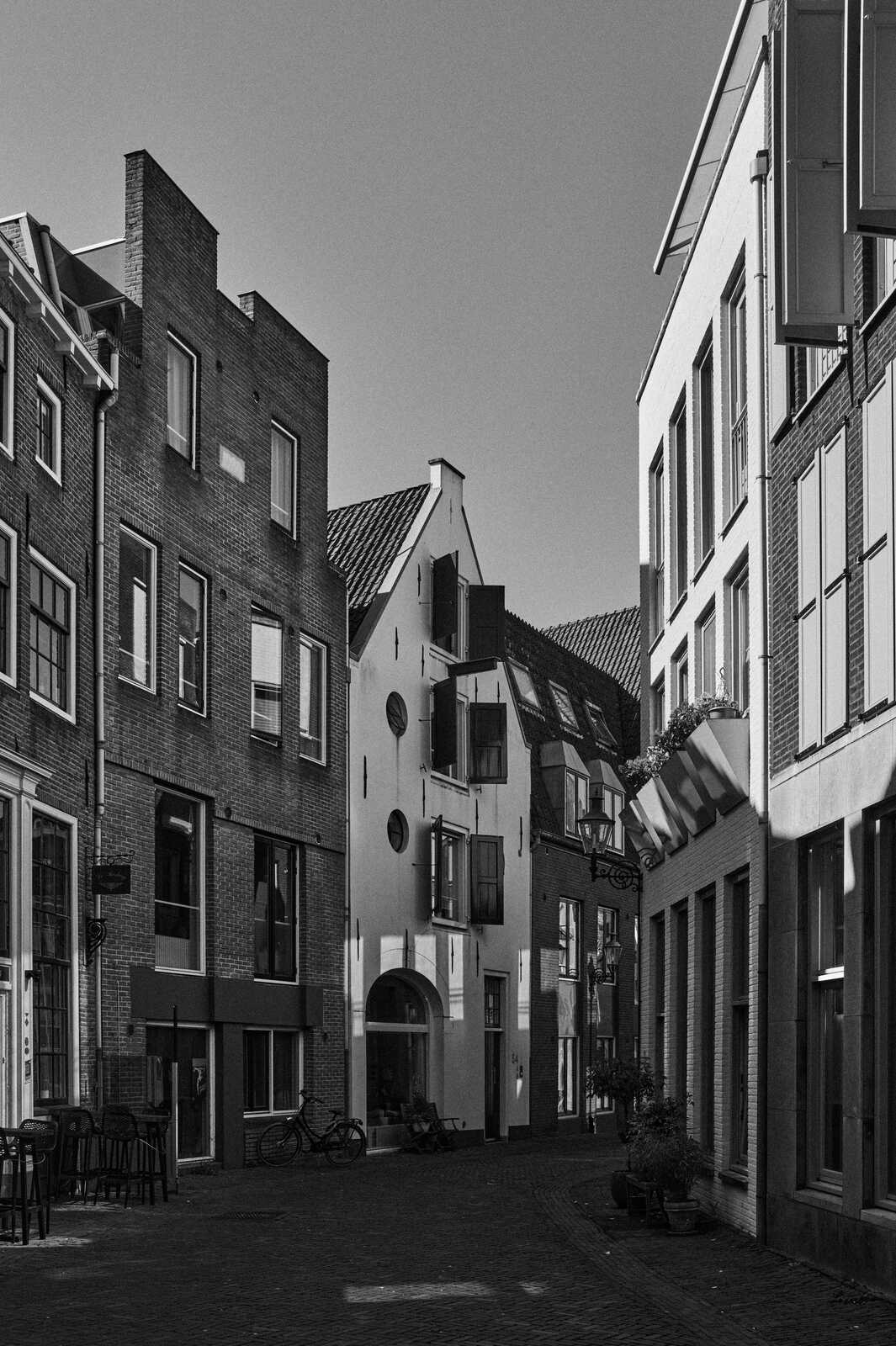 Black and white photo of narrow street lined with tall, gabled buildings. Cobblestone pavement, bicycles, and planters are visible. Several windows and doors are present.