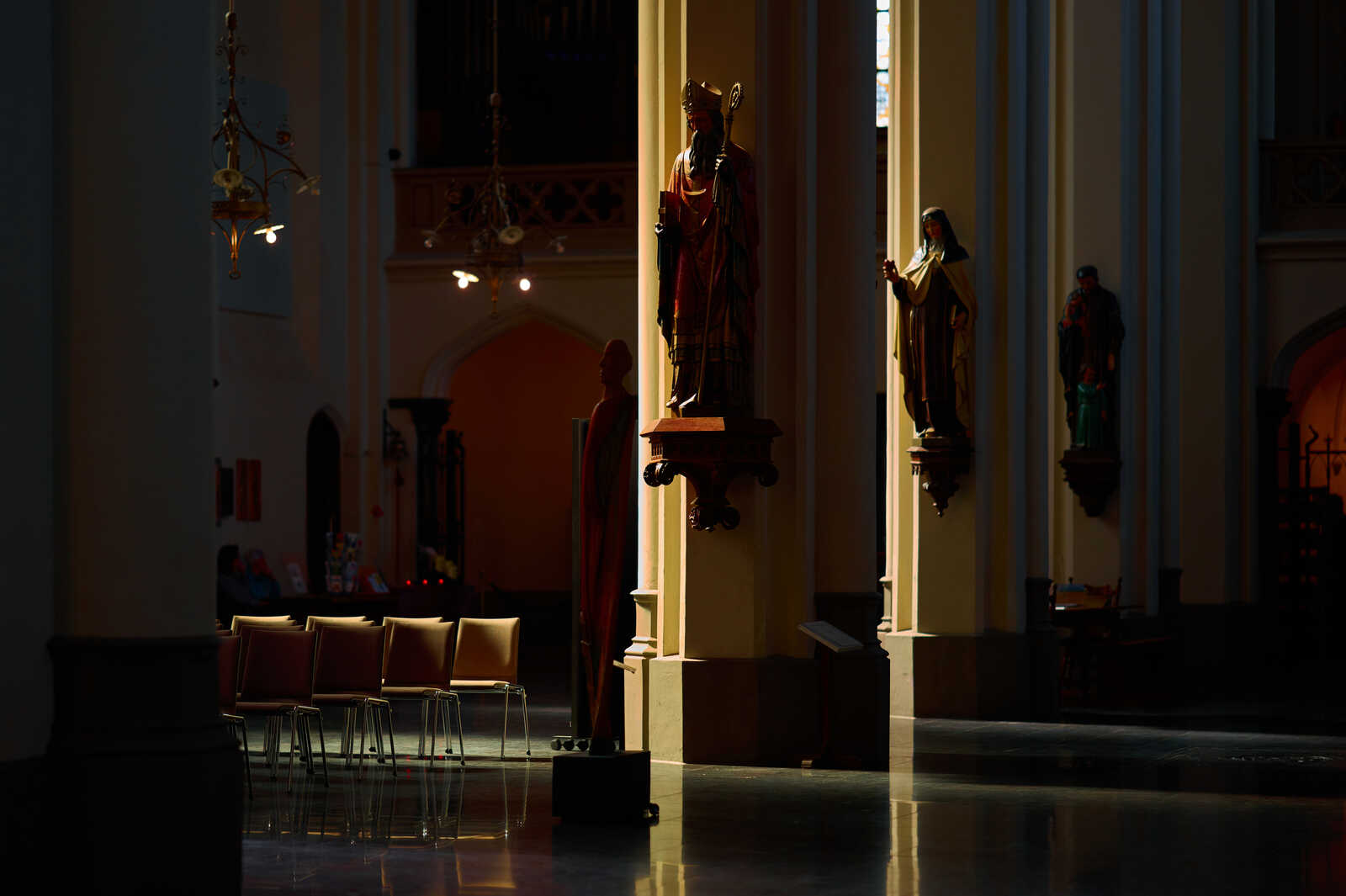 Interior of a church with two statues on pedestals. Dark pews are visible in the background, and a stained-glass window is partially seen. Soft, warm light illuminates the scene.