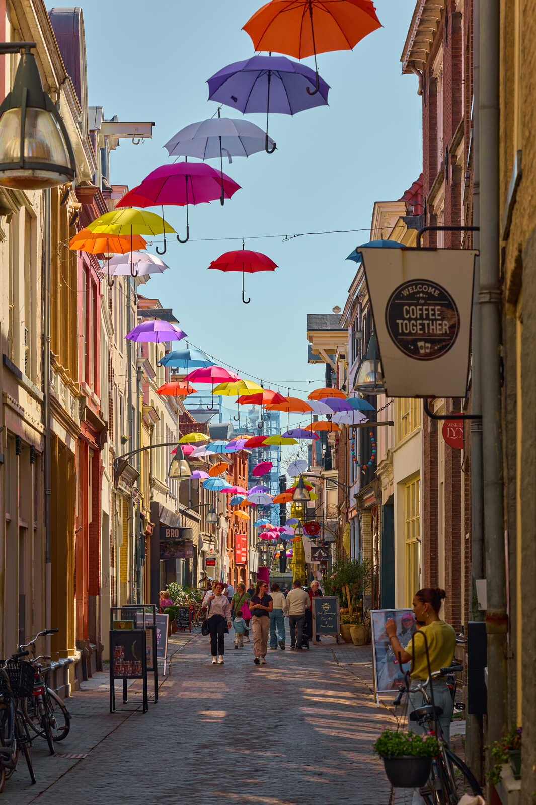 Colorful umbrellas hang above a pedestrian street lined with shops and cafes. People stroll along the walkway. A bicycle rests near a potted plant. Blue sky visible at the top.