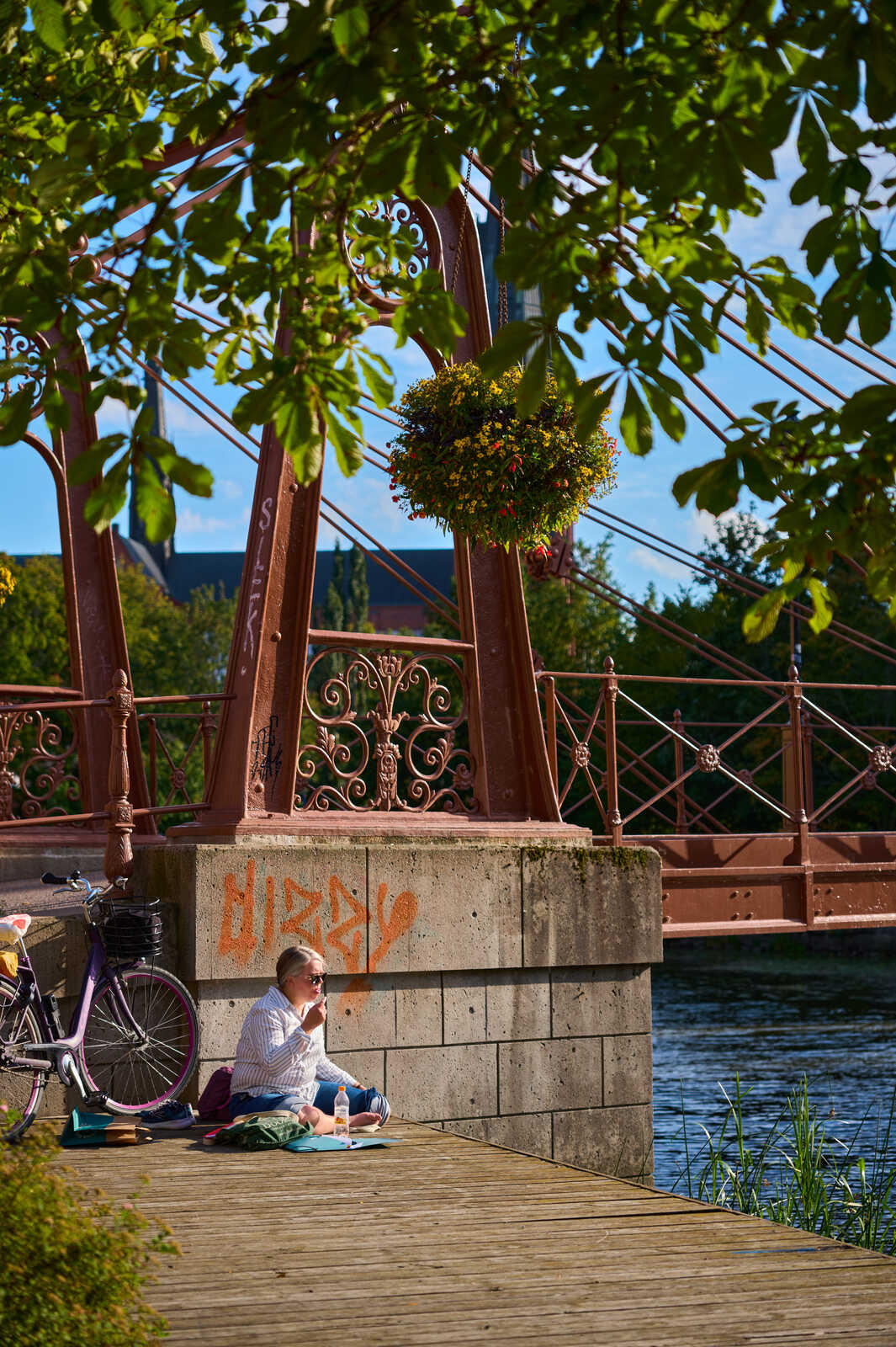 A woman sits on a wooden boardwalk near a bridge, reading a book. A bicycle is nearby. The bridge has red metalwork and a flower basket hangs from it. Water and greenery are visible behind the boardwalk.