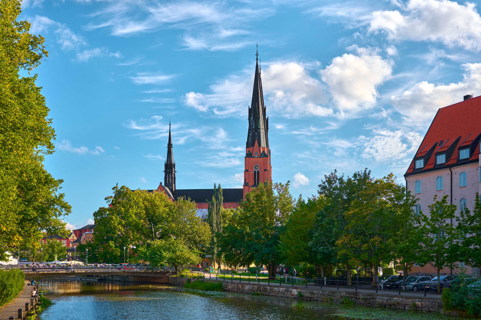 Tall church spire dominates the scene. A waterway with a bridge and greenery flanks it. Buildings with red and pink facades are visible alongside the riverbank under a partly cloudy blue sky. Cars and people are present.