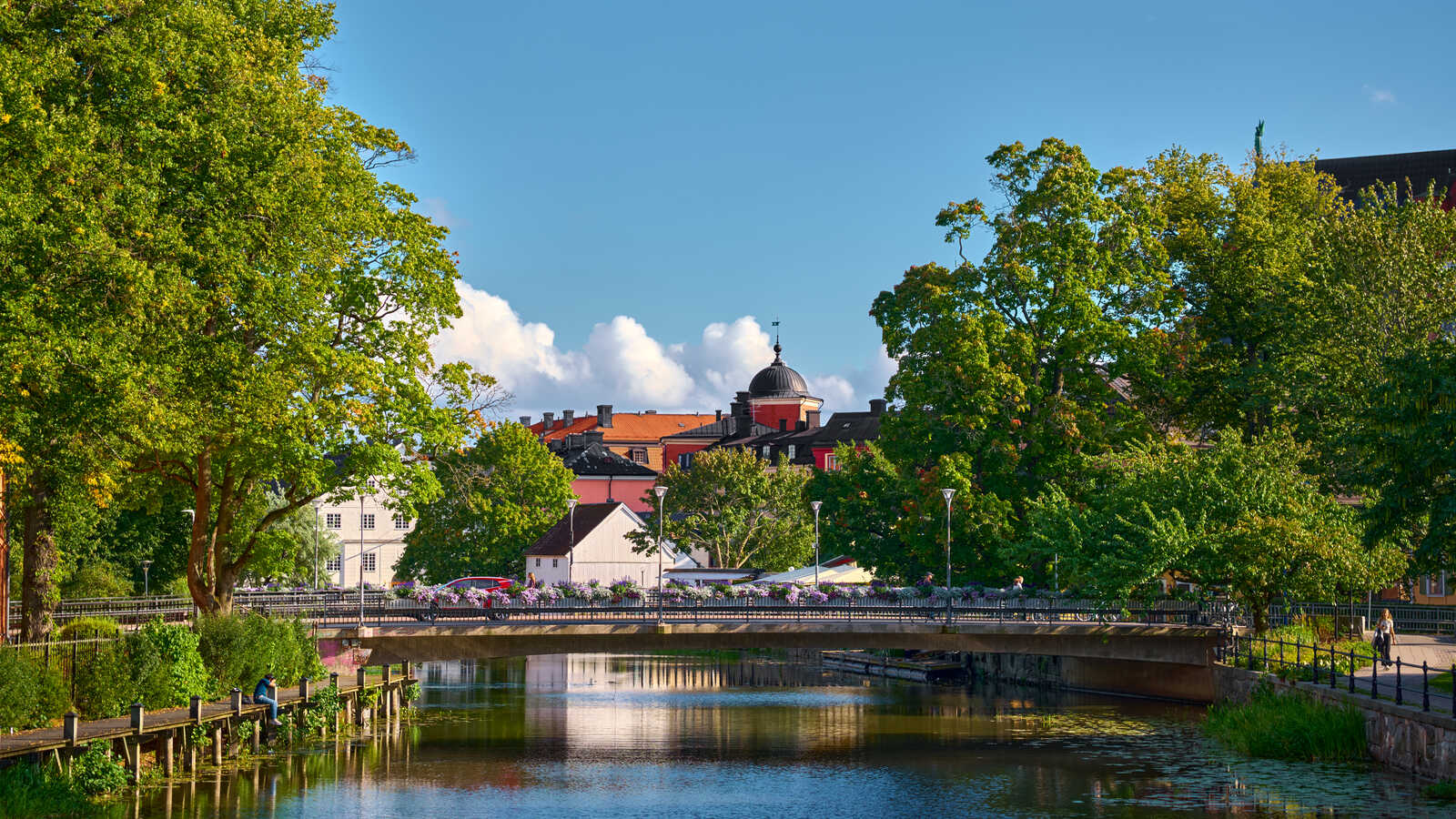 A river flows beneath a stone bridge lined with trees. Buildings with red and white facades are visible behind. A blue sky with fluffy white clouds is above. People are on the bridge and riverbank.