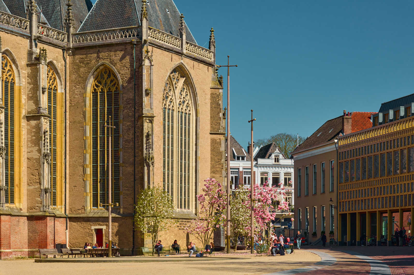 A large, ornate, golden-toned church with tall windows faces a sandy square. People relax on benches near a blooming tree and building with latticework details. Clear blue sky above.