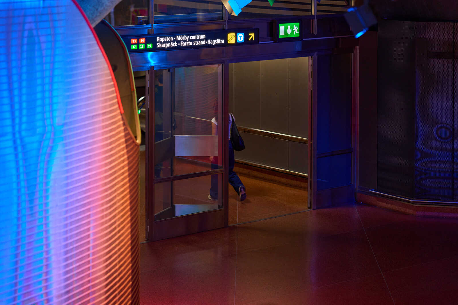 A glass door with a metal frame and signage. The background consists of a curved, illuminated red wall with text and a staircase.