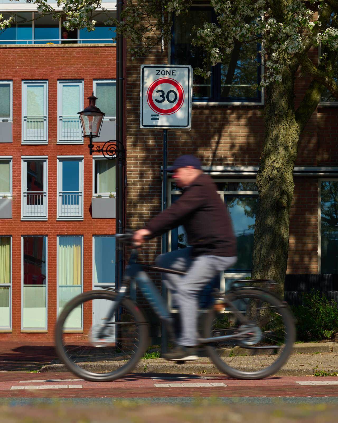 A person rides a bicycle on a street with a 30 speed limit sign. They wear a coat and helmet. A brick building and tree are in the background. The bike tires are black.