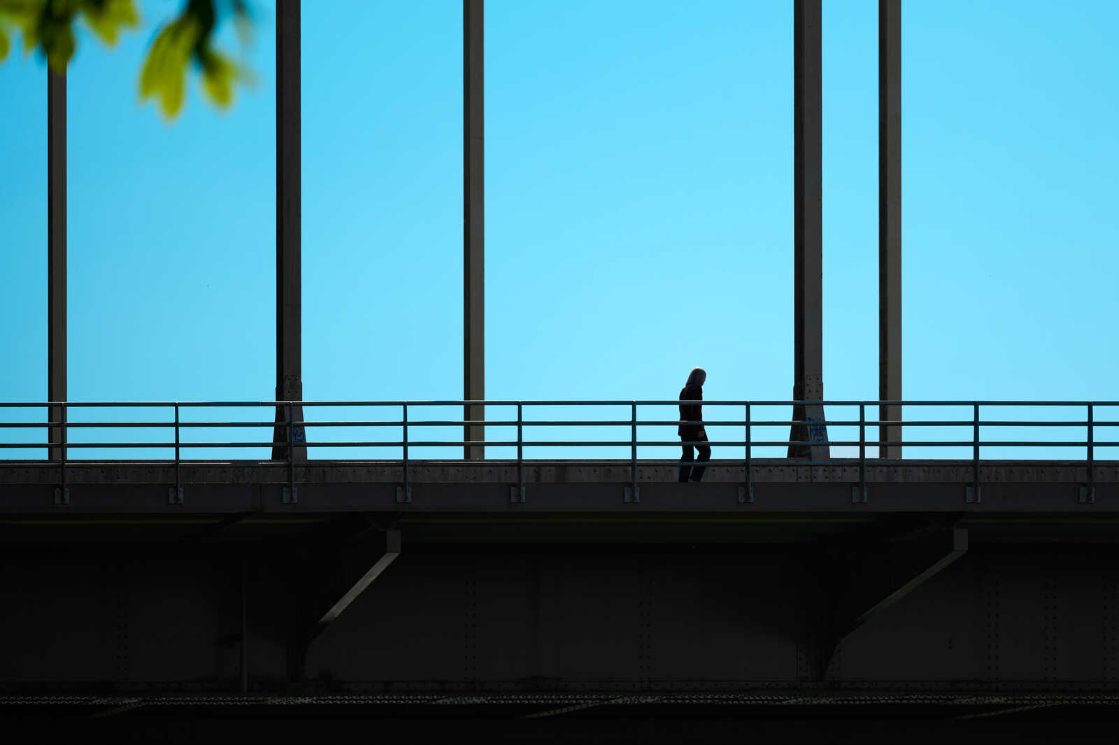 A silhouetted figure walks along a gray metal walkway with a railing against a bright blue sky. The walkway features dark structural supports.