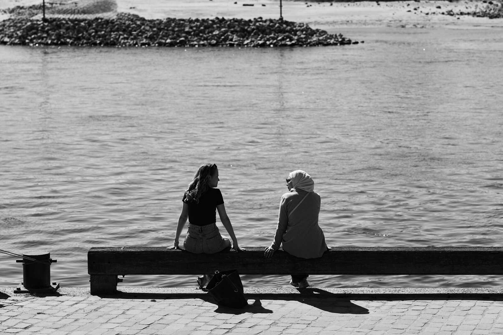 Two figures sit on a weathered wooden bench overlooking calm water. One wears a hijab, the other a black top and light-colored pants. A bag rests at their feet. The scene is monochrome.