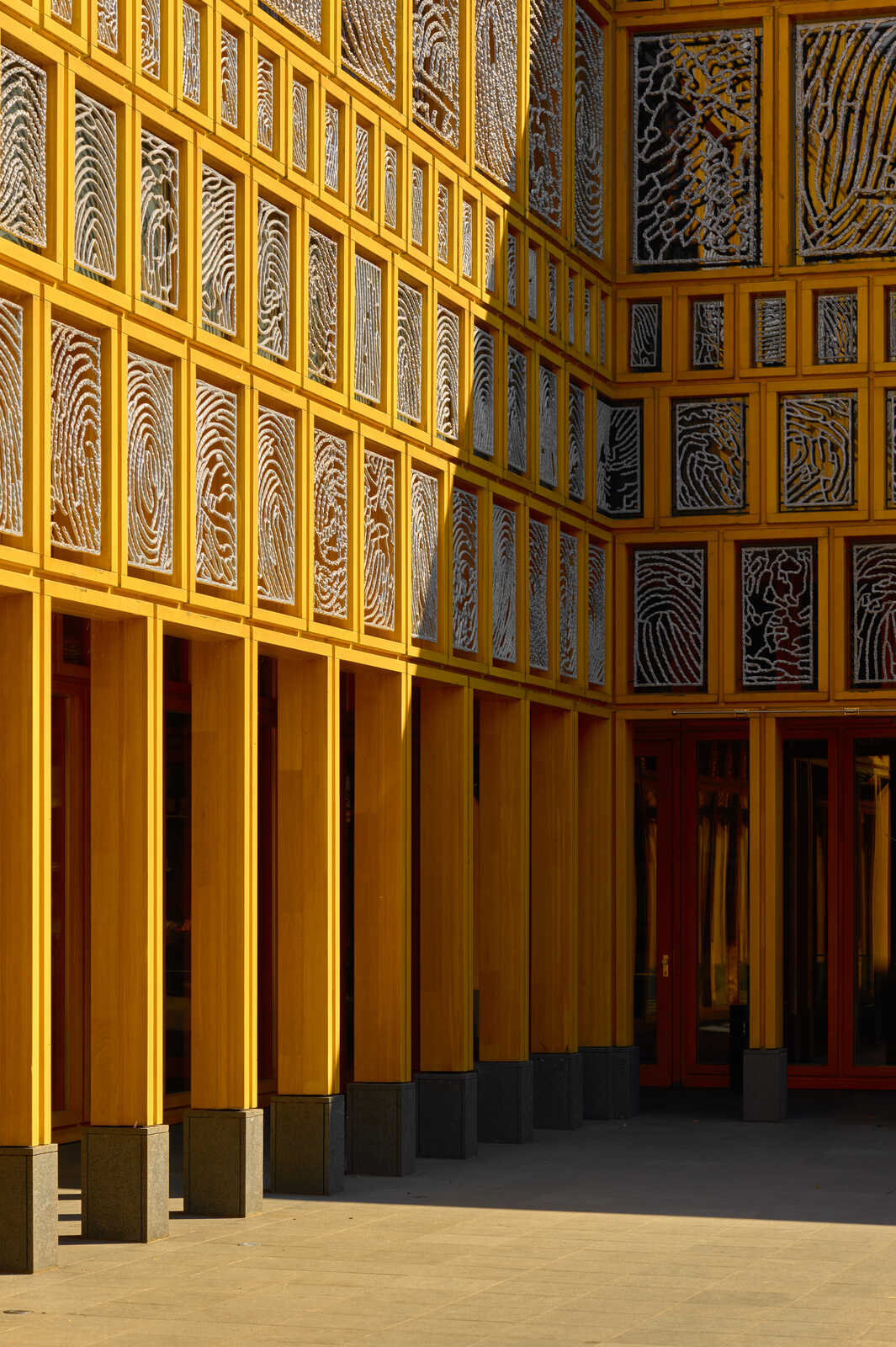 Yellow facade with patterned screens and red doors. Gray stone bases support pillars. Light casts shadows on a paved walkway. Modern architecture, bright and symmetrical.