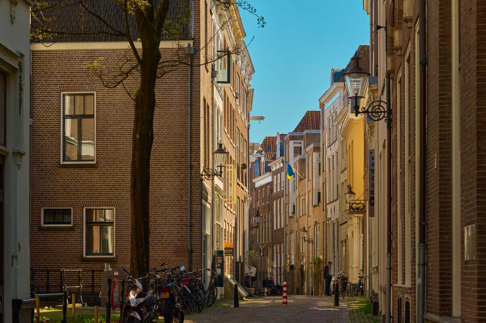 A cobblestone street lined with tall, colorful brick buildings. Bicycles are parked along the side. Street lamps and signage are visible. A person is walking further down the street. Clear blue sky above.