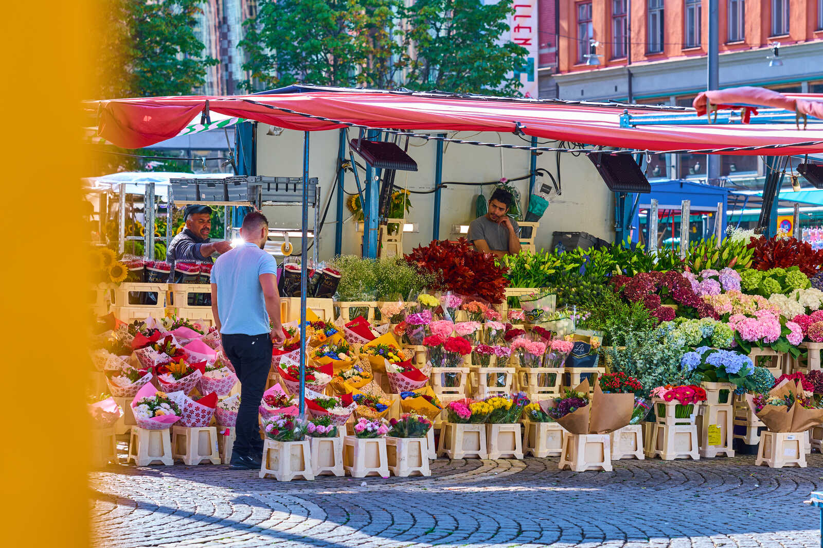 A man in a white shirt stands before a flower stand. Bouquets of various colorful flowers are displayed. A person is visible behind the counter. The scene takes place on a paved street. Red awnings cover the stand.