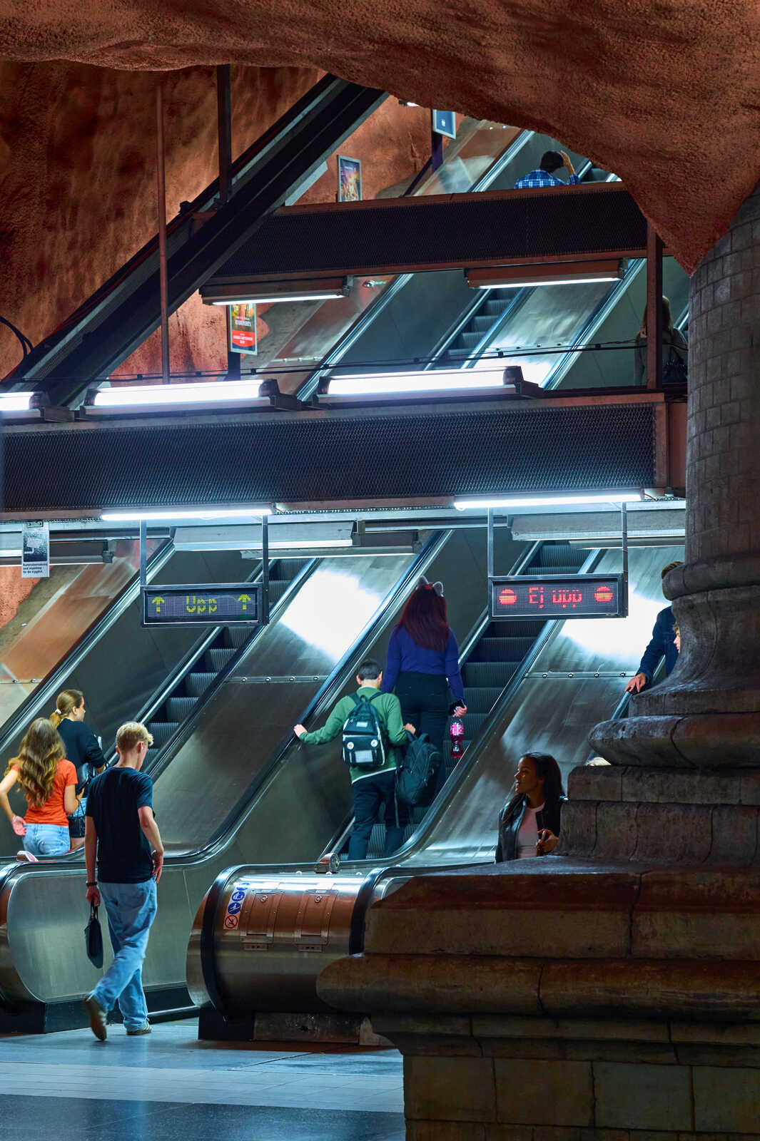 People ascend and descend on escalators within a stone and metal subway station. Digital signs display information. A concrete pillar and tiled wall are visible.