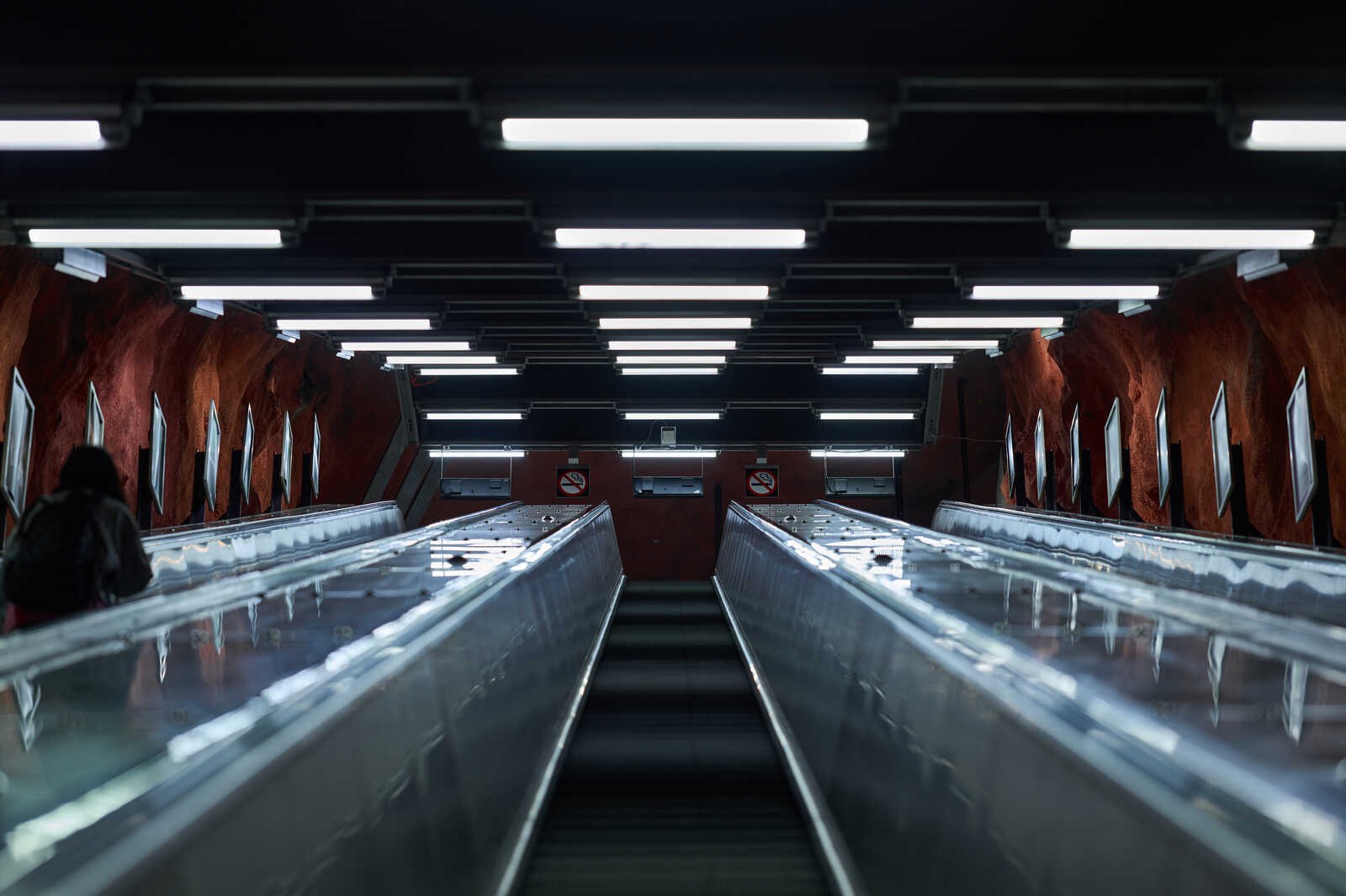Two escalators run parallel with metal railings. Overhead are numerous fluorescent lights against a red-brown wall. A person stands at the top, facing away. Signs are visible on the wall.