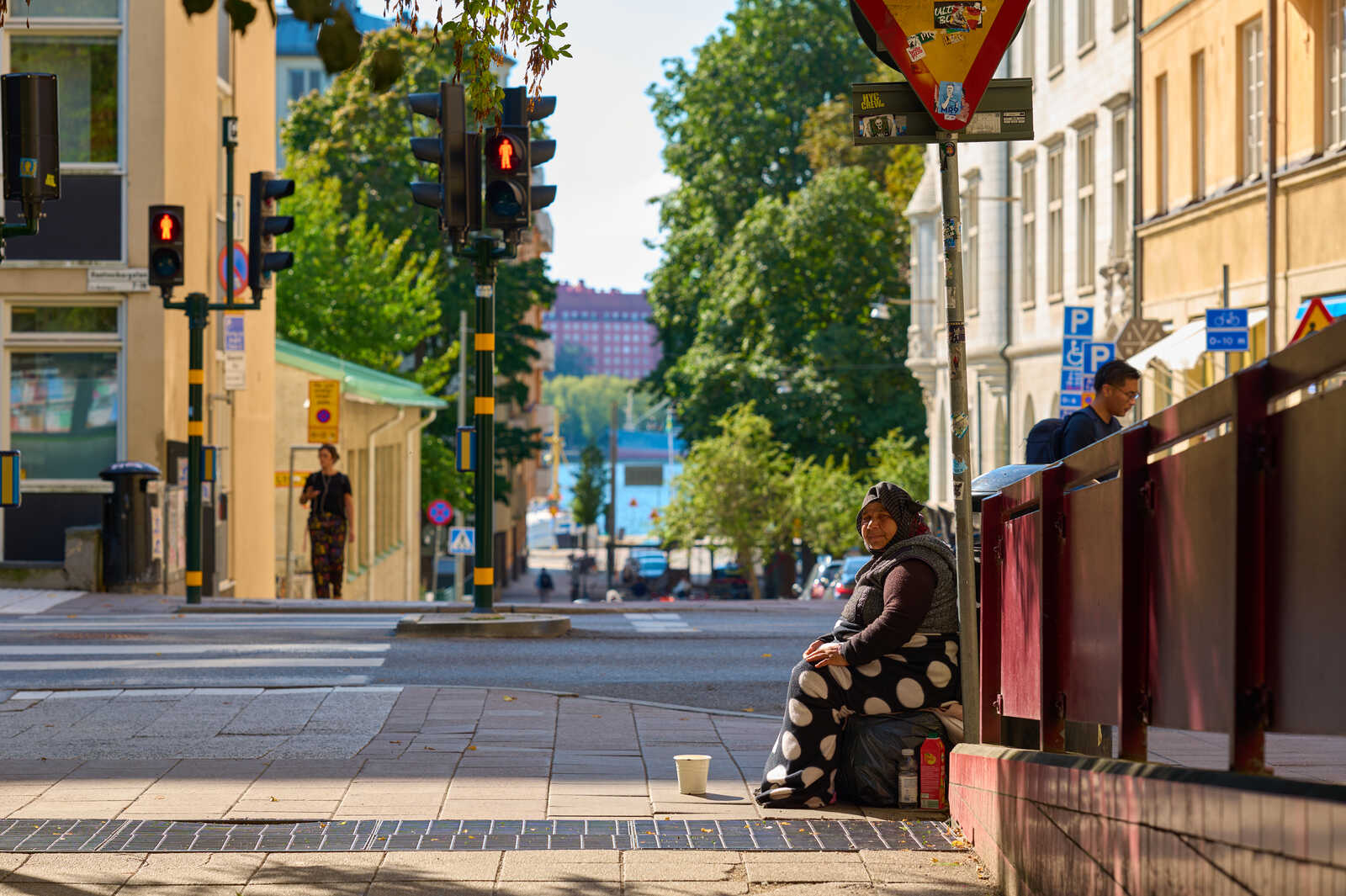 A woman sits on the sidewalk, wearing a patterned headscarf and dark dress. A cup sits before her. A street and building are behind. A person walks away in the distance.