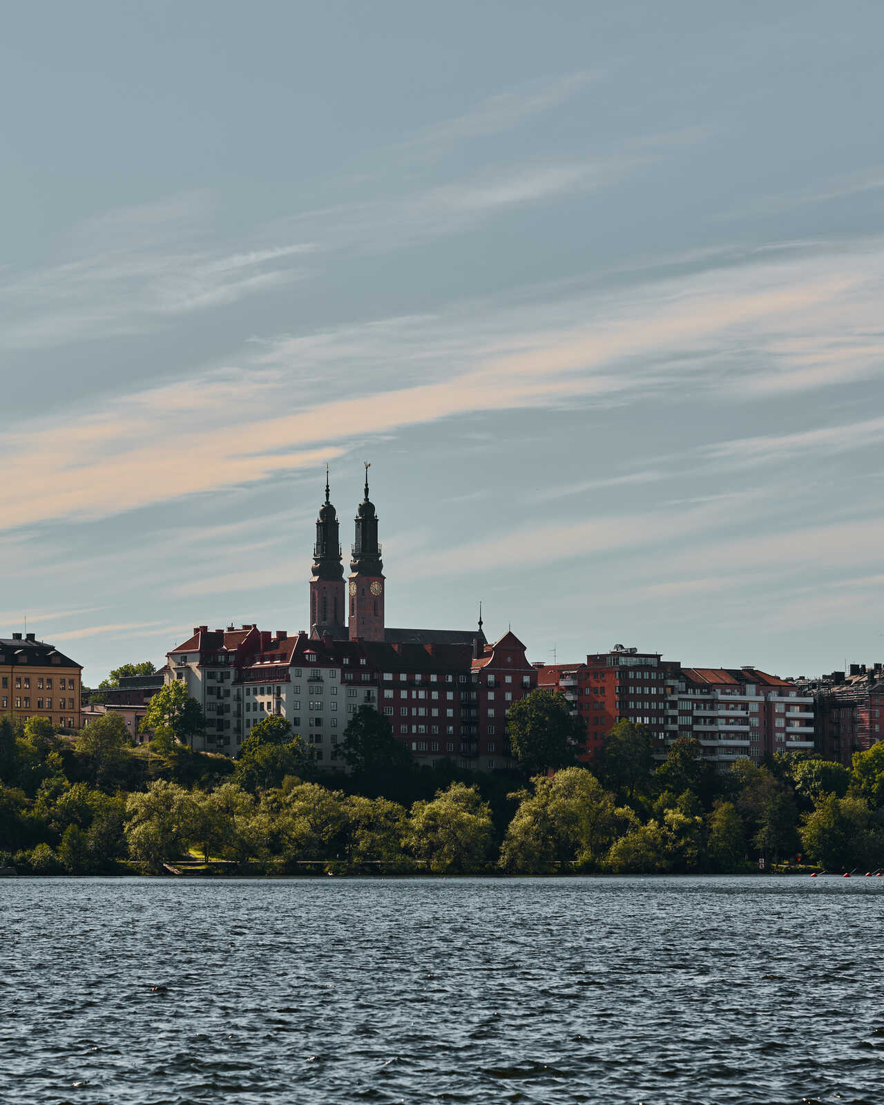 A city skyline with red-roofed buildings and a church with two towers is seen across a body of water. Trees line the shore, and a cloudy sky is above.