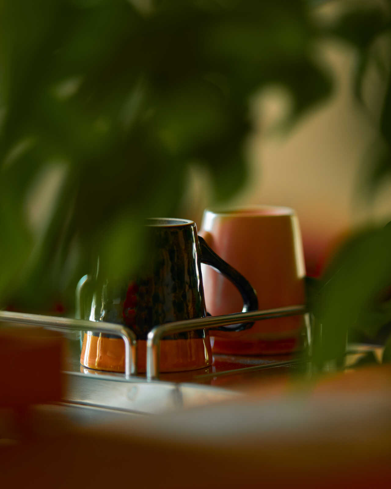 Three mugs sit on a silver tray. One mug is dark green with a handle, while the other two are a muted orange-red hue. The background is out of focus and blurred.