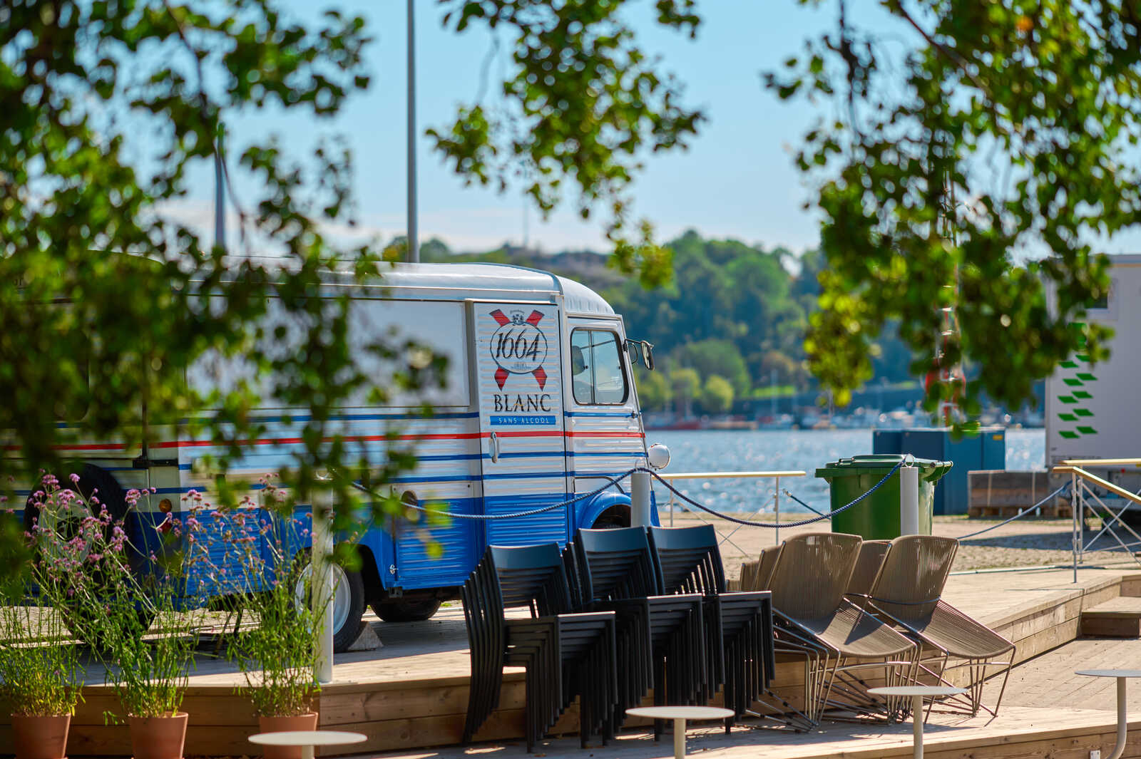 A vintage white Citroën HY van with blue trim stands on a wooden deck overlooking a lake. Several black chairs are stacked nearby. Green containers and trees are in the background. 100% Blanc is written on the van.