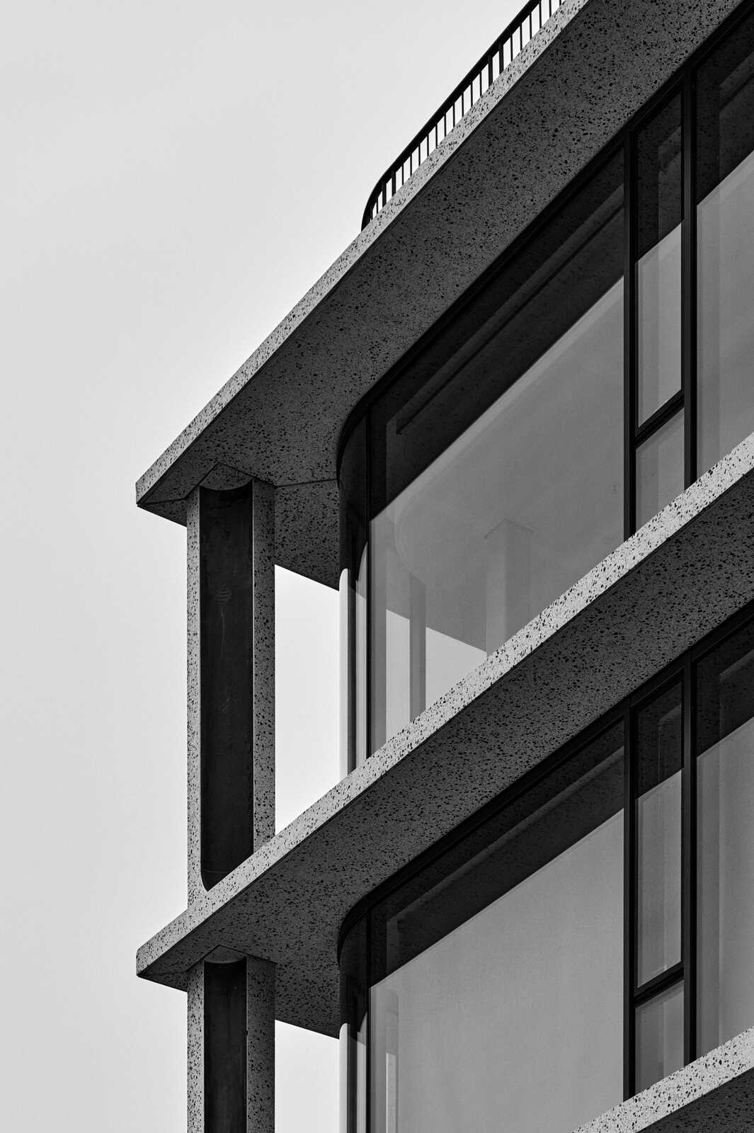 A grayscale image shows a corner of a modern building. It features textured concrete, large windows, and a slight architectural curve. A railing is visible at the top.