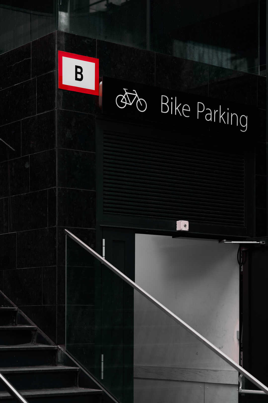 Black metal roll-up door with a red Bike Parking sign. Gray concrete steps with black railings lead upwards beside it. The wall is dark gray stone. Minimalist style.