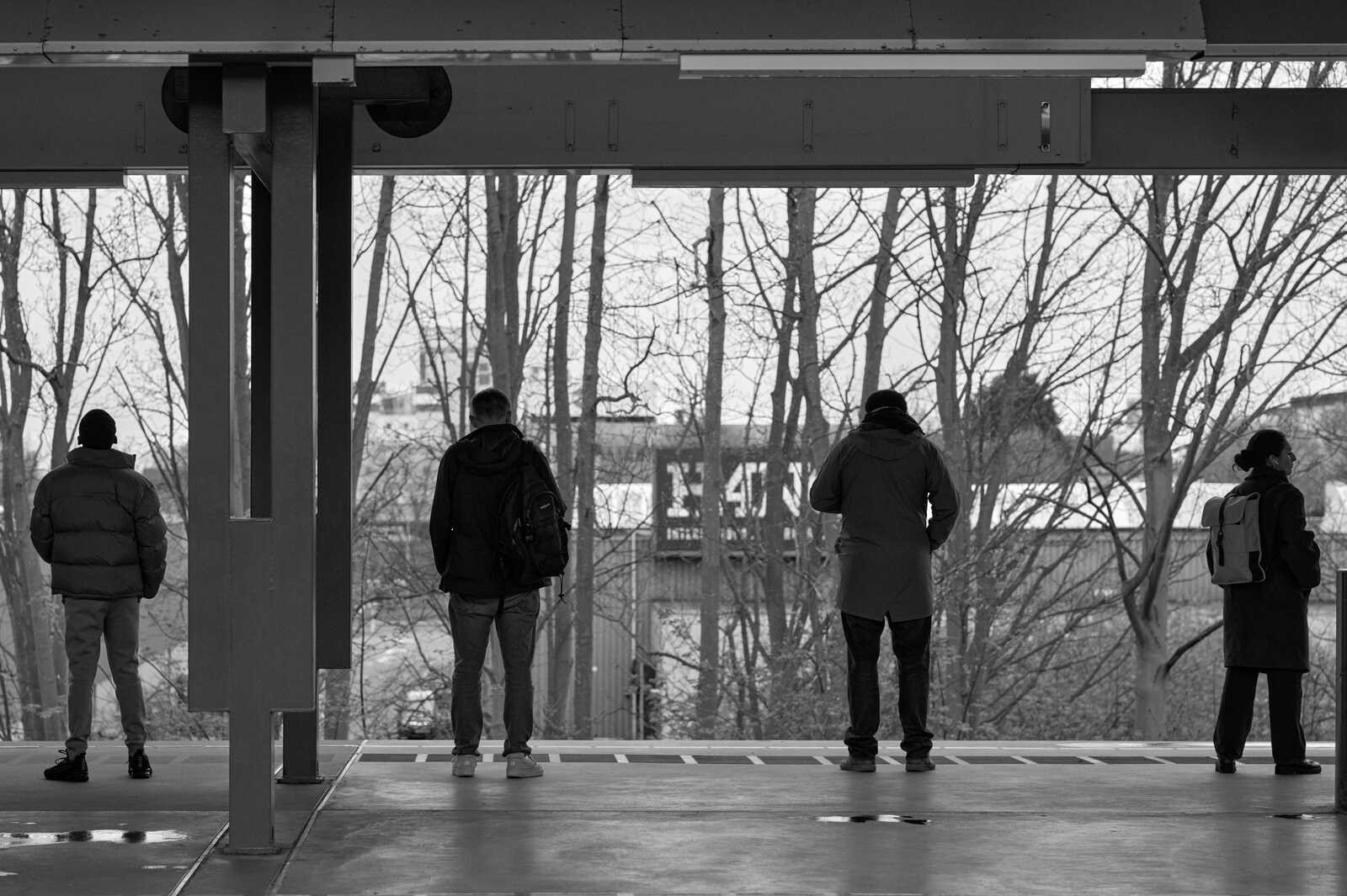 Four people stand on a platform, viewed from behind. They wear coats, jackets, and backpacks. A metal structure extends overhead. Trees are visible in the background.