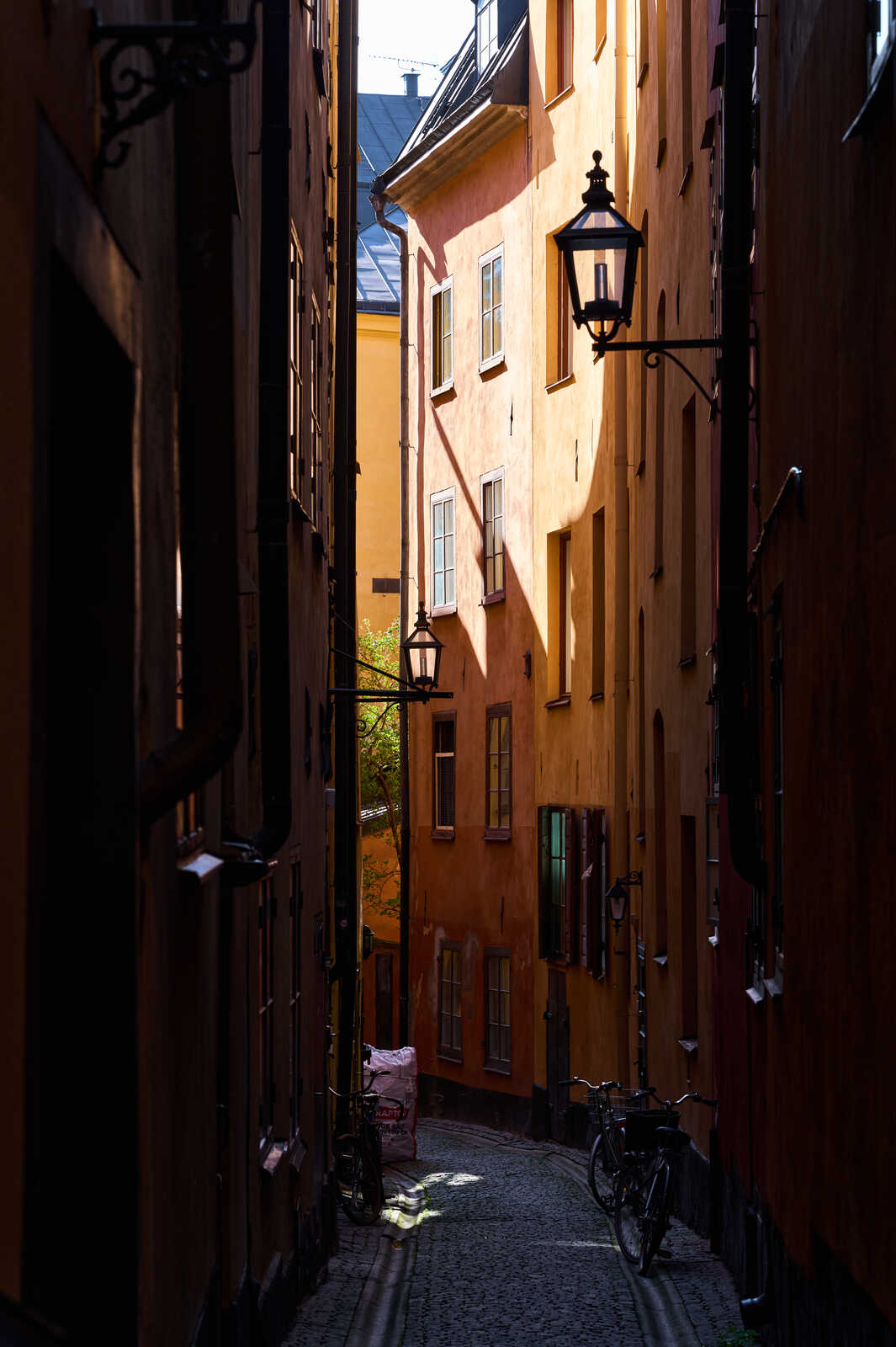 A narrow, cobblestone street with orange buildings. A black lamppost, bicycle, and a bag sit on the cobblestones. Green foliage peeks above the buildings. The street recedes into the distance.