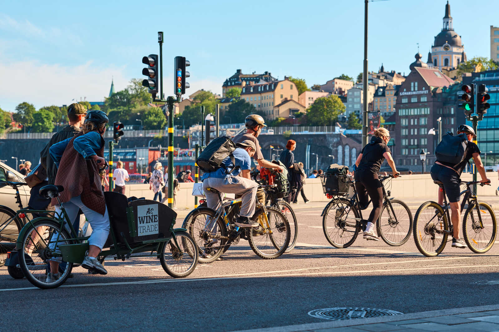 Four people on bicycles are crossing a street, including one on a large cargo bike with WINE TRADE on it. Buildings and a bridge are visible in the background. Traffic lights are present.