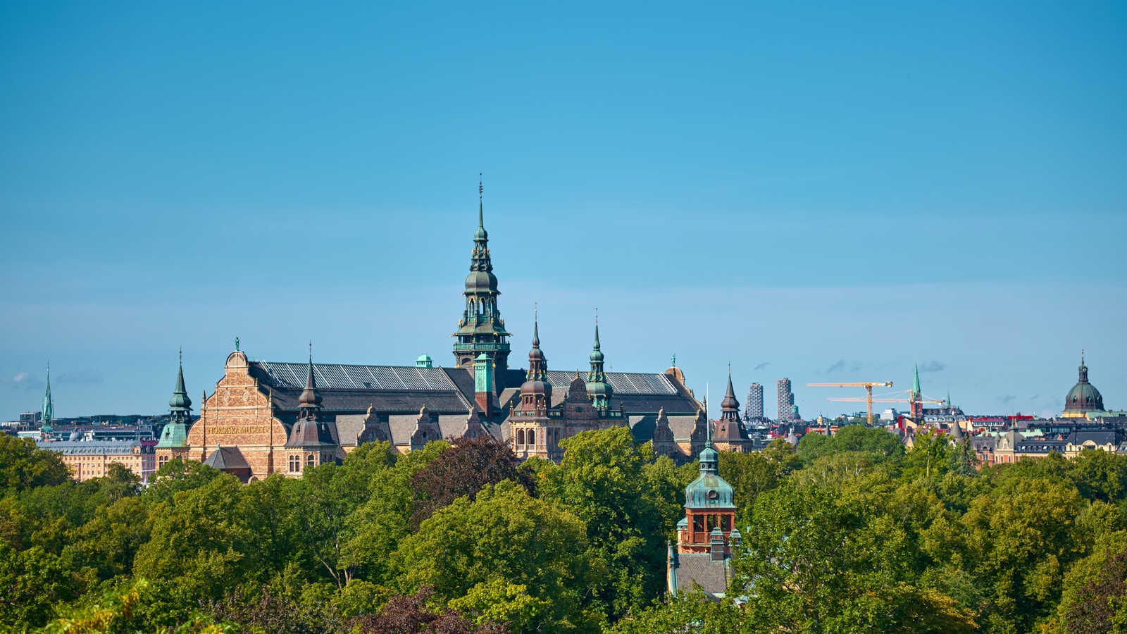 A high-angle shot shows a large, ornate building with multiple spires and a complex roofline, surrounded by lush green trees. A city skyline is visible in the background.
