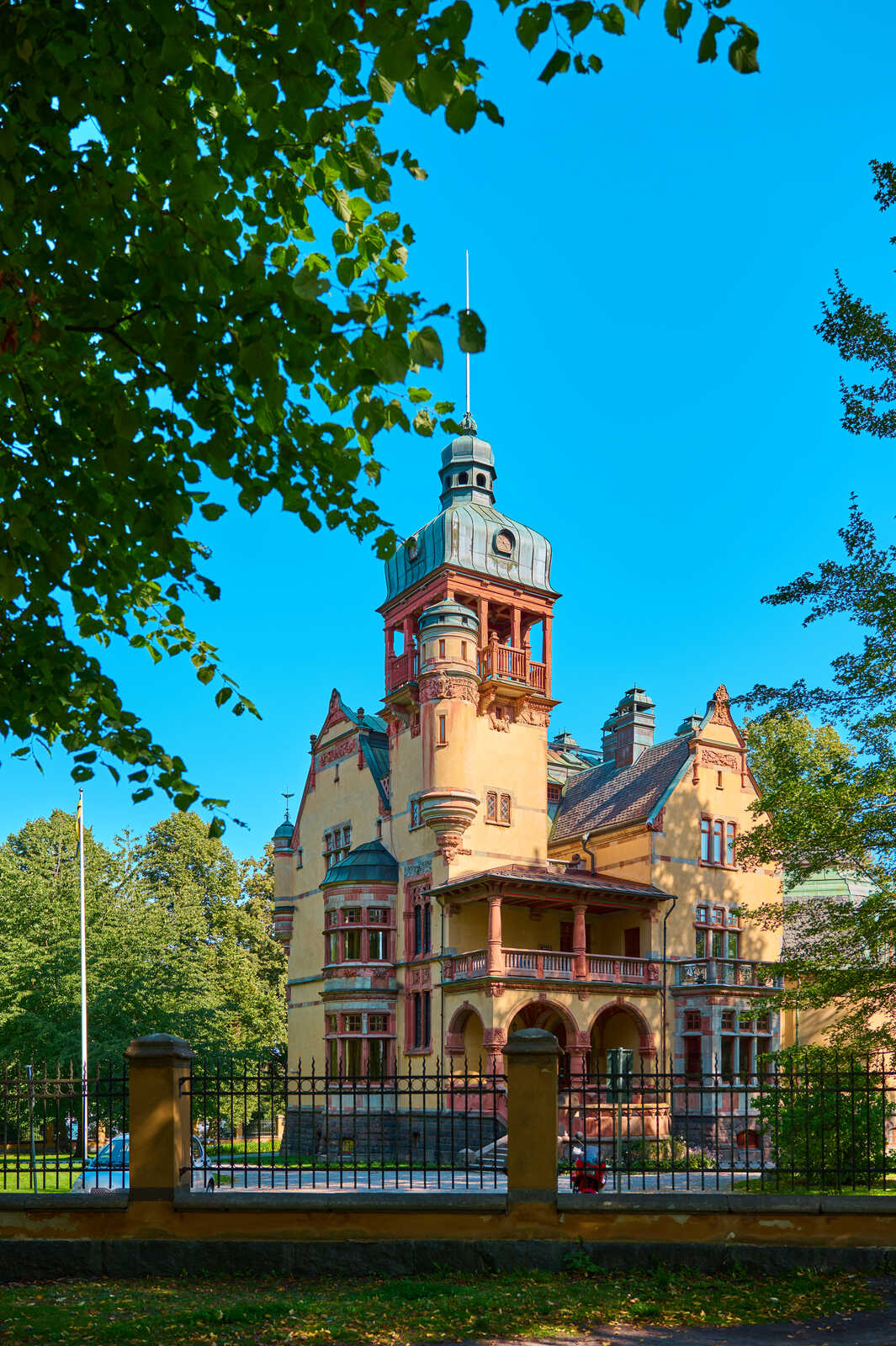 A large, ornate, two-story building with a central turret and multiple balconies, framed by a black wrought-iron fence and lush greenery under a clear blue sky. A flag pole stands nearby.