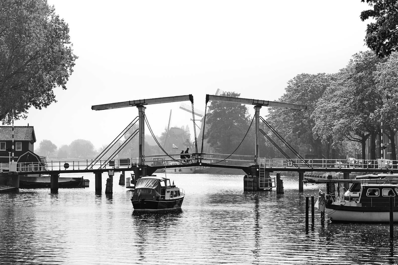A grayscale image shows a drawbridge over a canal. Two boats are moored alongside the bank. A windmill is visible in the distance under a cloudy sky. Tree foliage fills the left side.