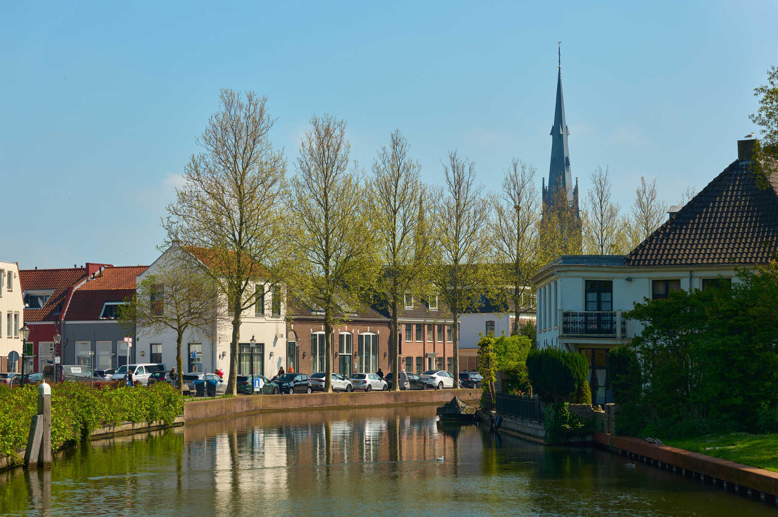 A canal with reflections of houses and trees. A church steeple rises in the background. Cars are parked along the canals edge, and greenery lines the banks. It’s a sunny day.