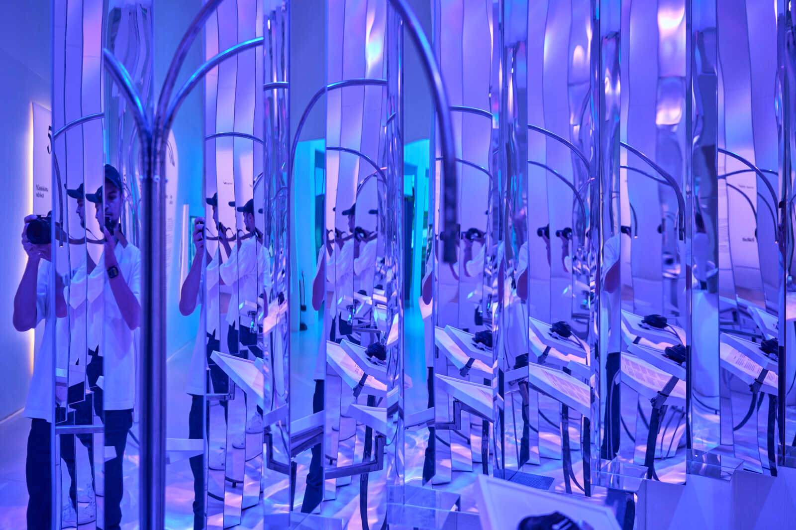 A person in a white t-shirt stands within an immersive, mirrored installation. Reflective panels and structures create a disorienting optical illusion. Blue lighting illuminates the scene. A camera is held in their hand.